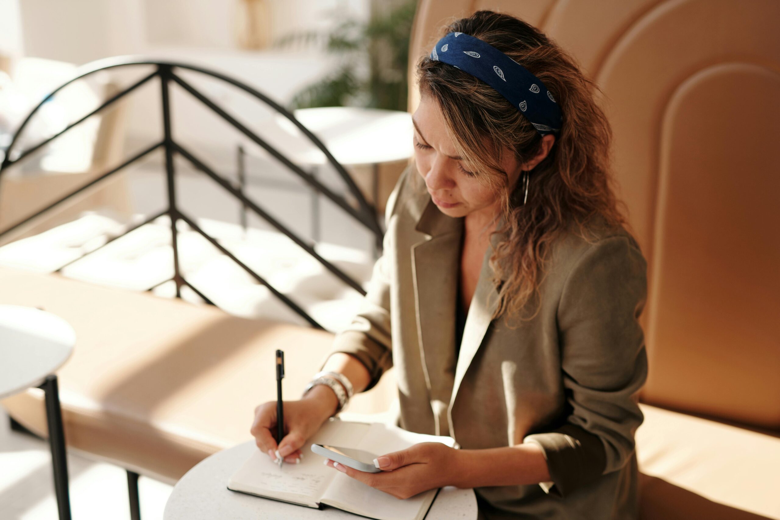 Woman in blazer writing notes while using smartphone in sunlit cafe.